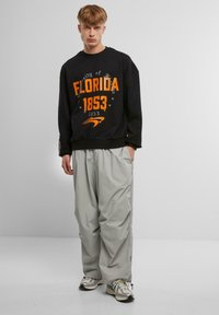 Fekete kapucnis szürke pulóver narancssárga "Florida Egyetem 1853" felirattal, világosszürke, bő szabású nadrággal és ezüstszínű sportcipővel párosítva.