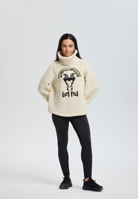 Cremfarbener Fleecepullover mit hohem Kragen und schwarzem Grafikdesign auf der Vorderseite, kombiniert mit schwarzen Leggings und Sportschuhen.