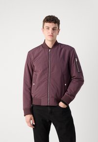 Maroon bomber jacket with a zip front, ribbed cuffs, and side pockets. Smooth texture and a zippered pocket on the sleeve. Black pants.