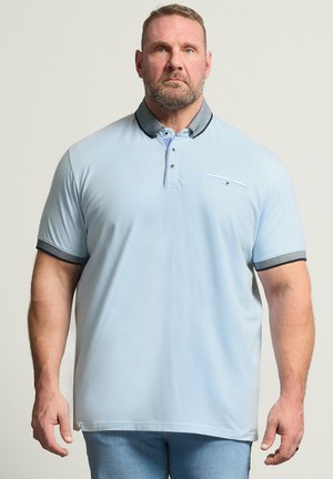 Dorosły mężczyzna z brodą, ubrany w jasnoniebieską koszulkę polo z ciemnym kołnierzykiem i mankietami, stojący na prostym jasnym tle.