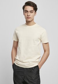 Licht beige katoenen T-shirt met een ronde hals en korte mouwen, gecombineerd met zwarte broek, weergegeven tegen een neutrale achtergrond.