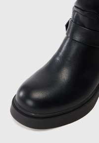 Primer plano de una bota de cuero negro con punta redondeada, detalle de correa y suela de goma gruesa sobre un fondo claro.