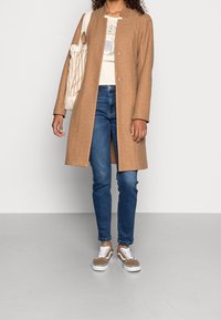Manteau en laine couleur fauve avec une rangée simple de boutons, associé à un t-shirt graphique et un jean bleu. Les accessoires comprennent un sac fourre-tout rayé et des baskets marron.
