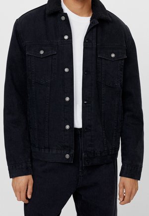 Homme portant une veste en jean noire avec poches boutonnées et col en shearling sur une chemise blanche, associée à un pantalon noir.