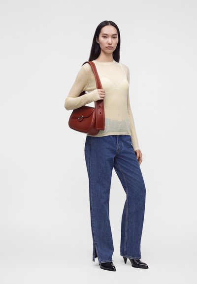 Femme portant un haut crème transparent à manches longues, un jean bleu, des bottes noires pointues, et portant un sac à bandoulière en cuir marron.