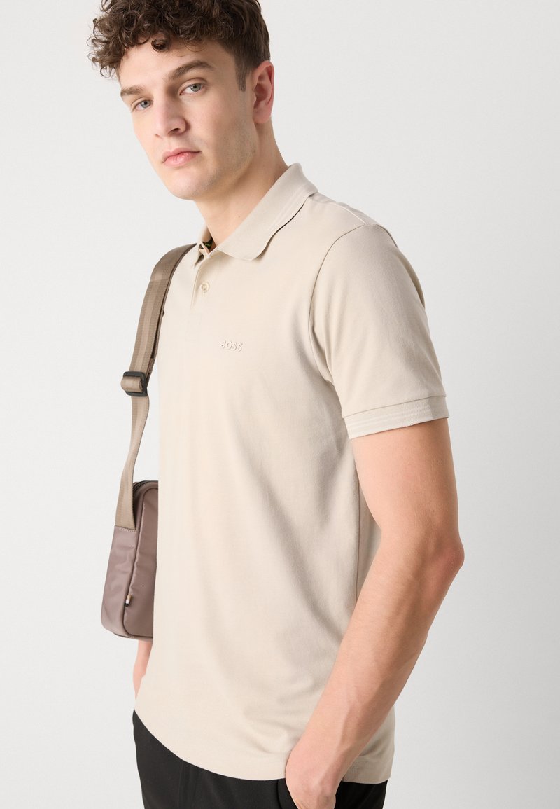 Polo shirt beige clair en coton doux, avec un col, une patte de boutonnage à deux boutons et un logo sur la poitrine. Sac messager marron sur l'épaule.
