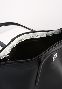 Musta synteettinen olkatasku, jossa on vetoketjullinen yläosa ja kudottu hihna, jossa lukee "LACOSTE". Edessä on pieni vihreä logo. Raaka pinta.