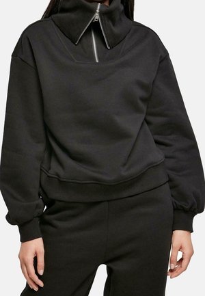 Nainen pukeutuneena mustaan pitkähihainen collegepaita, jossa on korkea kaulus ja hopeanvärinen vetoketju, yhdistettynä vastaaviin mustiin housuihin.