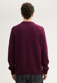 Pull en tricot bordeaux foncé avec un col rond, des poignets et un ourlet côtelés. Coutures apparentes au niveau des épaules. Coupe décontractée, tissu texturé.