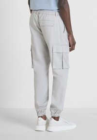 Pantalons cargo gris avec des poignets élastiques, des poches latérales et arrière, et une coupe ample. Portés avec des baskets blanches. Texture de tissu lisse.