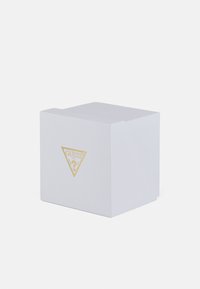Boîte texturée blanche avec un logo triangulaire doré GUESS à l'avant. Forme carrée avec un couvercle à rabat. Design simple et moderne.