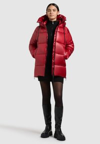 Rode gewatteerde parka met een ritssluiting aan de voorkant, hoge kraag en verstelbare capuchon. Heeft een glanzende textuur en gequilt detail. Draagt over een zwarte gebreide jurk.