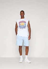 Vit ärmlös T-shirt med färggrafik "Dodgers World Series Champions"; ljusblå shorts; vita sneakers; neutral bakgrund.