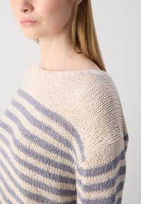 Femme portant un pull en maille à rayures crème et gris, aux cheveux longs et raides, tournée vers la gauche sur un fond blanc uni.