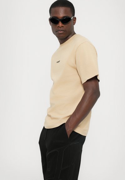 T-shirt en coton beige avec des manches courtes et un logo discret, associé à un pantalon noir avec un design texturé et des poches. Lunettes de soleil noires.