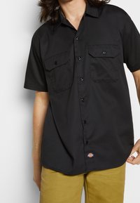 Černá košile na knoflíky s krátkým rukávem a dvěma náprsními kapsami, s nášivkou na lemu. Hladká látka a klasický design límce.