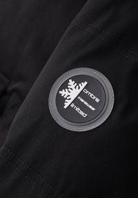 Tissu noir avec un écusson rond présentant un motif flocon de neige blanc et le texte "ombre menswear limited" en lettres blanches.