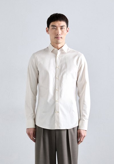 Chemise blanche à manches longues avec un motif rayé subtil, boutonnière et col pointu, portée avec un pantalon plissé gris.