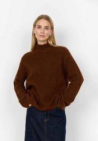 Brun, texturerad stickad tröja med hög krage och lös passform. Matchad med en mörk jeanskjol. Tyget verkar mjukt och varmt.