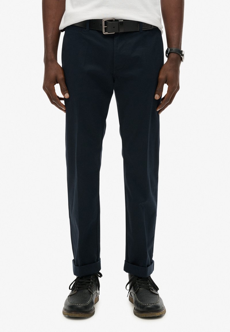 Navy Chinos mit geradem Schnitt, umgeschlagenen Säumen und glatter Textur, kombiniert mit einem schwarzen Gürtel und dunklen Schuhen. Minimalistische Details.