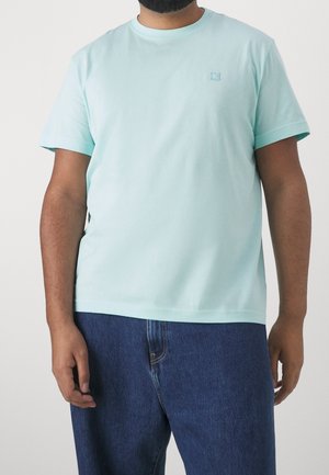 Homme portant un T-shirt à manches courtes bleu clair et un jean en denim bleu, debout devant un fond clair uni.