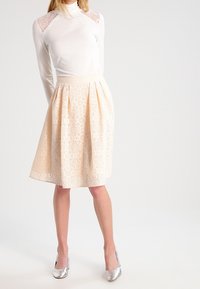 Valkoinen ribattu poolopaita, jossa on pitsiset olkaimet, yhdistettynä polven pituiseen, laskostettuun vaaleanbeigeen hameeseen, jossa on pitsi päällä, ja hopeisiin korkokenkiin.