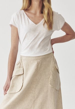 Mujer con una camiseta blanca de manga corta y escote en V, metida dentro de una falda beige de lino con grandes bolsillos delanteros, de pie con una mano en la cadera.