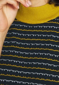 Stickad tröja med en ribbad senapsgul krage, som har växelvis randiga mönster i mörk marinblått, gult och vitt med en texturerad yta.