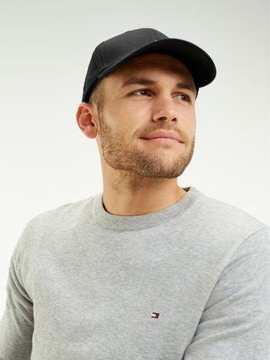 Junger Mann mit kurzem Bart, der eine schwarze Mütze und einen hellgrauen Sweatshirt mit kleinem Logo trägt, und leicht nach oben und zur Seite schaut.