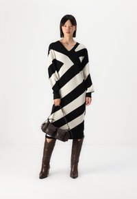 Robe en tricot avec un motif zigzag noir et blanc, décolleté en V, manches longues. Associée à un sac à main marron foncé et des bottes marron jusqu'aux genoux.