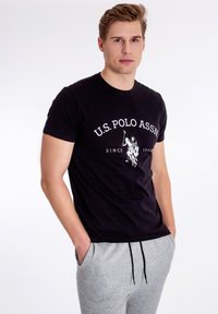 Sort bomulds T-shirt med en hvid grafisk print af "U.S. Polo Assn." og en polo spiller. Sammen med grå sweatpants, hvilket viser et afslappet look.