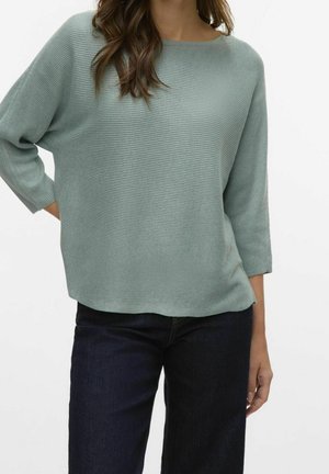 Femme portant un pull côtelé vert clair à manches longues et un jean bleu foncé, debout avec une main sur la hanche, fond blanc.
