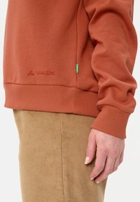 Sweatshirt couleur rouille avec des poignets et un ourlet côtelés. Présente un logo "VAUDE" ton sur ton et une petite étiquette verte. Texture douce, design décontracté.