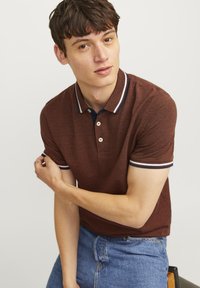 Bruine polo met een gestructureerde stof, voorzien van een marineblauwe kraag en gestreepte accenten op de mouwen. Knopen zichtbaar op de sluiting.