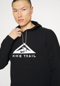 Musta huppari, jossa on kolmion muotoinen logo ja valkoiset "NIKE TRAIL" -tekstit. Siinä on teksturoitu pinta ja erottuva kengännauhatyyppinen nyöri.