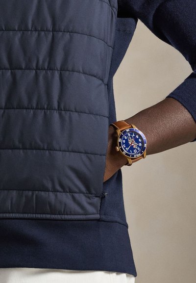 Doudoune sans manches bleu marine à motif matelassé diagonal à côté d'une montre en or avec un cadran bleu, un bracelet en cuir marron et de grands chiffres blancs.