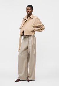 Γυναίκα που φοράει ένα μπεζ cropped σακάκι με φαρδιές πέτες και μπεζ ψηλόμεσα, φαρδιά παντελόνια με ζώνη, στέκεται μπροστά σε μονόχρωμο φόντο.