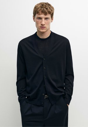 Giovane uomo con capelli corti e ricci che indossa un cardigan nero abbottonato sopra una camicia nera con le mani in tasca contro uno sfondo semplice.