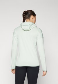 Lys grøn hettegenser med lange ermer, figursydd design, strukturert stoff og sidelommer. Båret over svarte bukser.