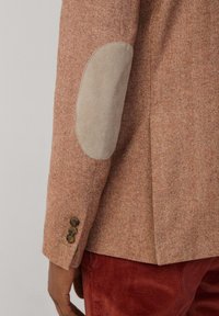 Braune Jacke im Fischgrätmuster mit beigen Ellbogenpatches und drei bronzenen Knöpfen am Ärmel. Strukturiertes Material mit taillierter Passform.