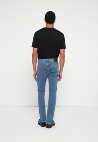 Must t-särk ja helesinised teksapüksid tagantvaates, millel on vöökohal nahast plaastriga detail ja kitsas lõige, kantud mustade kingadega.