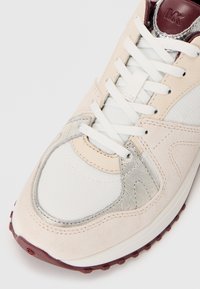 Sneaker beige e bianco con lacci bianchi, dettagli argentati e suola bordeaux, in primo piano che mostra la punta e la parte superiore.