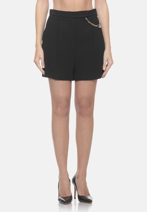 Pantaloncini neri su misura con un piccolo dettaglio di catena dorata indossati con scarpe nere con tacco, mostrati dalla metà del torso fino ai piedi.