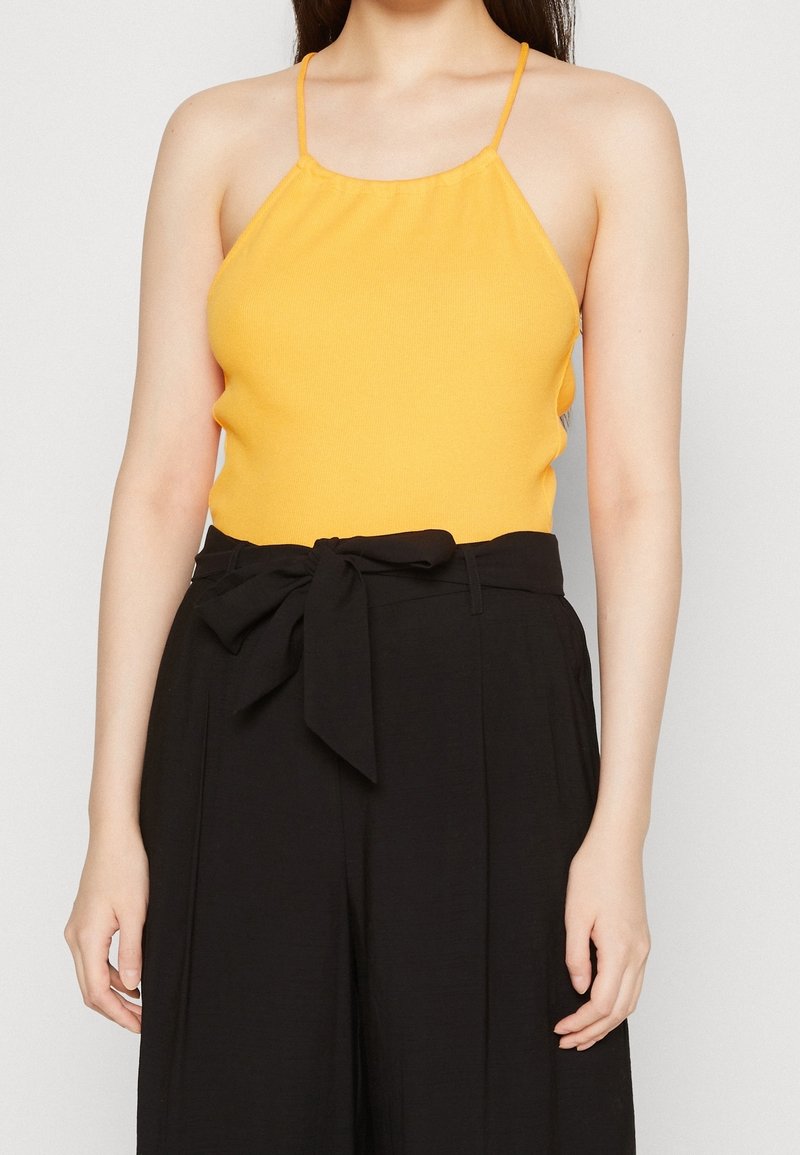 Femme portant un haut jaune sans manches avec de fines bretelles et un pantalon noir taille haute noué à la taille avec une large ceinture en tissu.