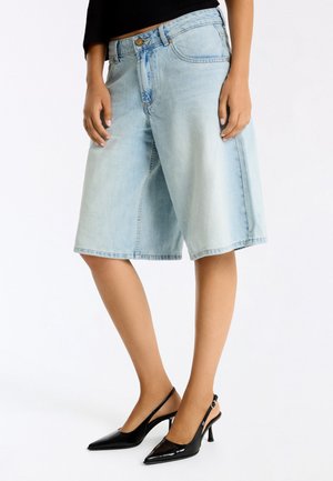 Person iført knælange lyseblå denimshorts og sorte spidse slingback-højhælede sko, stående med armene afslappet ned langs siderne.
