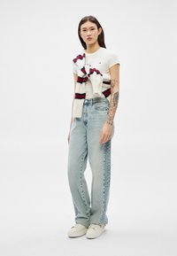 Sieviete ar gariem tumšiem matiem, valkā baltu t-kreklu, gaiši zilas džinsa bikses, baltas sporta kurpes un baltu džemperi ar sarkaniem un melniem svītrām, kas pārmests pār pleciem.