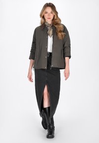 Olivengrüne, leichte Jacke mit Druckknopfverschlüssen, kombiniert mit einem schwarzen Denim-Rock mit hohem Schlitz. Das Model trägt kniehohe schwarze Stiefel und einen Schal.