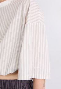Top corto a maniche corte di colore beige chiaro con righe bianche verticali. Presenta un piccolo logo Nike bianco sull'orlo. Tessuto morbido, vestibilità rilassata.