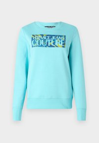 Világoskék pamutszvetter, amelyen téglalap alakú grafika található, rajta a "VERSACE JEANS COUTURE" felirat vastag fehér betűkkel, és színes, mintás háttérrel.