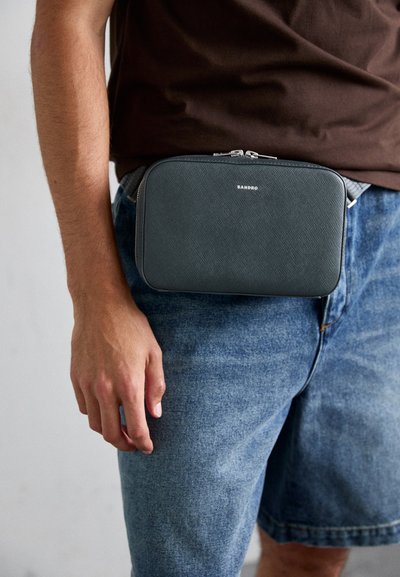 Borsa a pochette nera con texture, dotata di una cerniera argento, indossata in vita, con un logo semplice sul davanti, abbinata a pantaloni corti in denim.